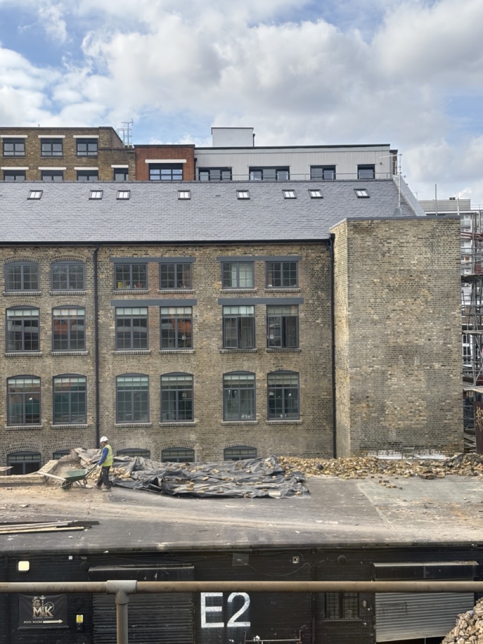 Photograph of a worker with a wheelbarrow next to a pile of bricks and black plastic sheeting on top of a rooftop in Bethnal Green, London. Behind a large brick building is shown